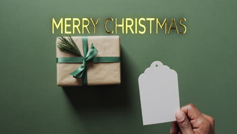 Frohe-Weihnachten-Text-In-Gold-Mit-Geschenk-Und-Afrikanischer-Amerikanischer-Männlicher-Hand-Mit-Leerem-Etikett-Auf-Grün
