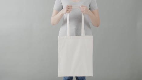 Mujer-Caucásica-Con-Camiseta-Blanca-Sosteniendo-Una-Bolsa-Blanca-Sobre-Fondo-Gris,-Espacio-Para-Copiar,-Cámara-Lenta