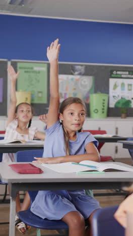 Vertical-video-of-happy-diverse-schoolchildren-raising-hands-in-classroom-in-slow-motion