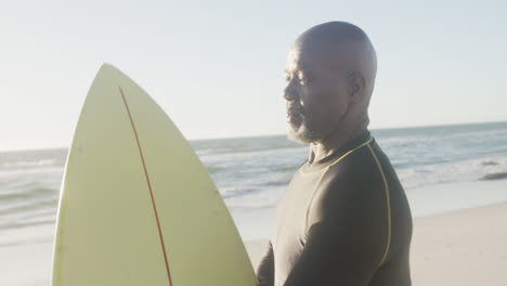 Happy-senior-african-american-man-holding-surfboard-at-beach,-in-slow-motion
