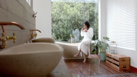 Biracial-woman-wearing-bathrobe-sitting-on-bathtub-drinking-tea-at-home,-slow-motion,-copy-space