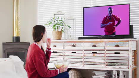 Caucasian-woman-watching-tv-with-african-american-male-rugby-player-catching-ball-on-screen