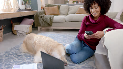 Mujer-Birracial-Feliz-Con-Perro-Golden-Retriever-Usando-Laptop-Y-Teléfono-Inteligente-En-Casa,-Cámara-Lenta