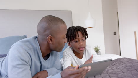 Afroamerikanischer-Vater-Und-Sohn-Liegen-Zu-Hause-Mit-Tablet-Im-Bett,-Zeitlupe