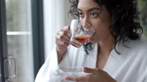 Mujer-Birracial-Usando-Albornoz-Bebiendo-Té-Y-Mirando-Por-La-Ventana-En-Casa,-Cámara-Lenta