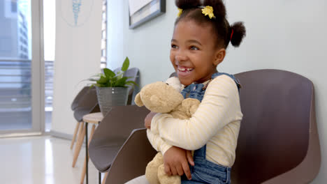 Happy-african-american-girl-sitting-in-hospital-waiting-room-and-hugging-mascot,-slow-motion