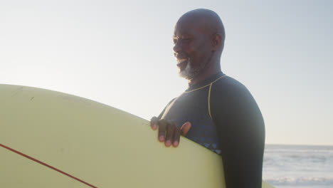 Glücklicher-älterer-Afroamerikanischer-Mann,-Der-In-Zeitlupe-Am-Strand-Spazieren-Geht-Und-Ein-Surfbrett-Hält