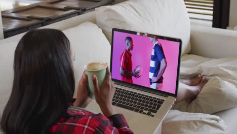 Biracial-woman-watching-laptop-with-diverse-male-rugby-player-with-ball-on-screen