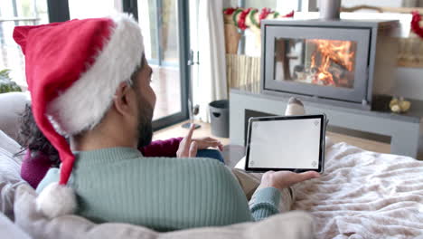 Biracial-couple-wearing-santa-claus-hats-using-tablet-with-copy-space-on-screen-at-home,-slow-motion