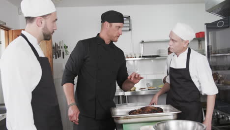 Chef-Masculino-Diverso-Enfocado-Instruyendo-A-Chefs-Masculinos-En-Formación-En-La-Cocina,-Cámara-Lenta