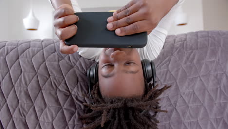 Glücklicher-Afroamerikanischer-Junge-Mit-Kopfhörern,-Der-Zu-Hause-Mit-Dem-Smartphone-Auf-Dem-Bett-Liegt,-Zeitlupe