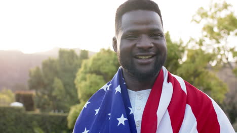 Glücklicher-Afroamerikanischer-Mann,-Der-Mit-Flagge-Auf-Dem-Rücken-Steht-Und-Im-Sonnigen-Garten-Lächelt