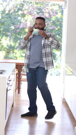 Vertical-video-of-african-american-man-drinking-coffee-talking-on-smartphone-in-kitchen,-slow-motion