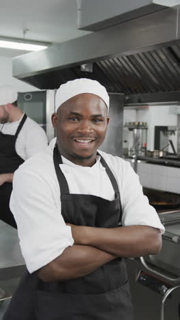 Happy-african-american-male-chef-standing-with-arms-crossed-in-kitchen,-slow-motion,-vertical