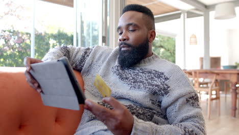 Glücklicher-Afroamerikanischer-Mann-Mit-Tablet-Und-Kreditkarte-Im-Sonnigen-Wohnzimmer,-Zeitlupe