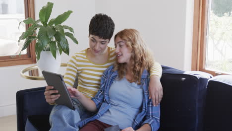 Happy-caucasian-lesbian-couple-embracing-on-sofa,-using-tablet-and-credit-card-in-sunny-house