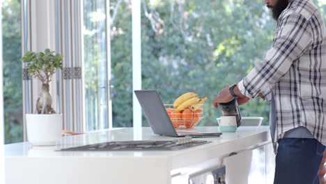 Hombre-Afroamericano-Haciendo-Café-Y-Usando-Una-Computadora-Portátil-En-Una-Cocina-Soleada,-Cámara-Lenta