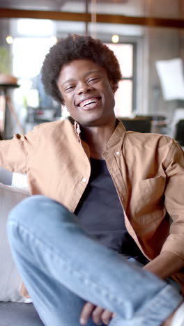 Vídeo-Vertical-Del-Retrato-De-Un-Feliz-Hombre-Afroamericano-Sentado-En-Un-Sofá-En-Casa,-Cámara-Lenta