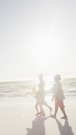 Verical-video-of-african-american-grandmother,-mother-and-daughter-walking-at-beach,-in-slow-motion
