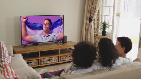 Biracial-family-watching-tv-with-caucasian-male-supporter-with-flag-on-screen