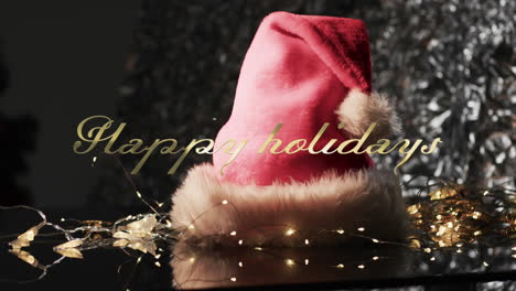 Happy-holidays-text-in-gold-over-christmas-hat,-lights-and-tree-on-dark-background