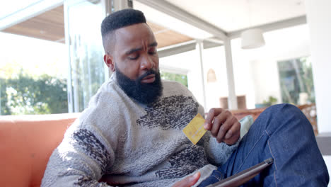 Hombre-Afroamericano-Usando-Tableta-Y-Tarjeta-De-Crédito-En-Una-Sala-De-Estar-Soleada,-Cámara-Lenta