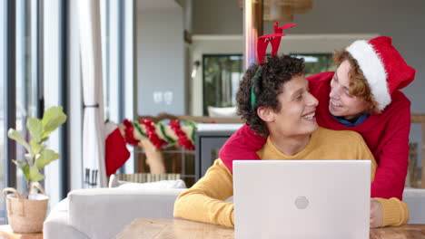 Diverse-gay-male-couple-using-laptop-for-video-call-at-home-at-christmas,-slow-motion,-copy-space