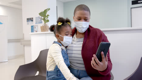 Afroamerikanische-Mutter-Und-Tochter-Tragen-Gesichtsmasken-Mit-Smartphone-Im-Krankenhaus,-Zeitlupe