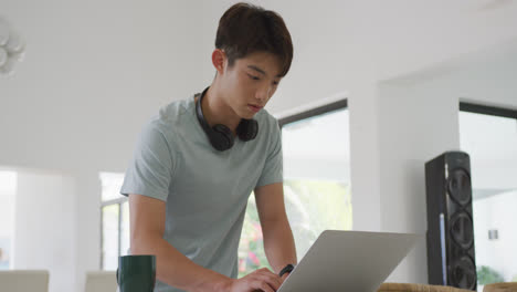 Asian-male-teenager-using-laptop-and-wearing-headphones-in-living-room