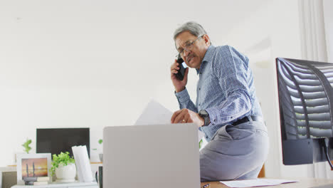 Video-Eines-älteren-Mannes-Mit-Gemischter-Abstammung,-Der-Am-Schreibtisch-Sitzt,-Telefoniert-Und-Zu-Hause-Seinen-Laptop-Benutzt