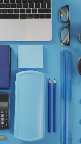 Vertical-video-of-laptop,-glasses,-coffee,-plant-and-stationery-on-blue-background