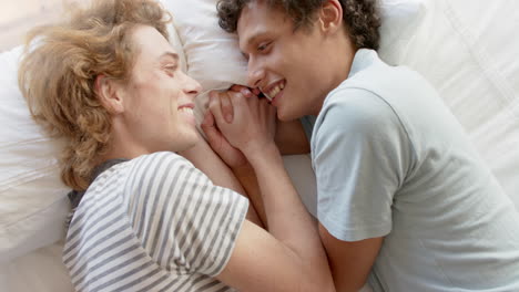 Happy-diverse-gay-male-couple-lying-on-bed-holding-hands-at-home,-slow-motion