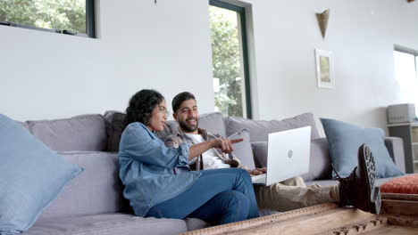 Happy-biracial-couple-sitting-on-sofa-and-using-laptop-at-home,-in-slow-motion