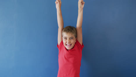 Portrait-of-happy-caucasian-boy-raising-hands-and-smiling-on-blue-background,-slow-motion