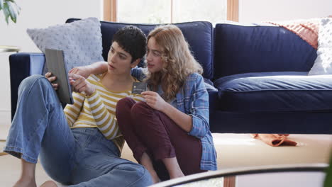 Happy-caucasian-lesbian-couple-embracing-on-floor,-using-tablet-and-credit-card-in-sunny-house
