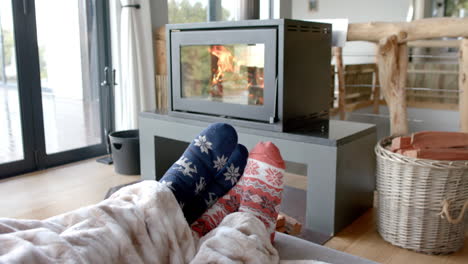 Feet-of-couple-on-sofa-in-warm-socks-under-blanket-in-front-of-fireplace-at-home,-in-slow-motion
