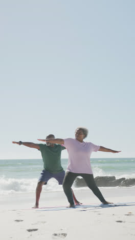 Vertical-video-of-senior-african-american-couple-doing-yoga-at-beach,-in-slow-motion