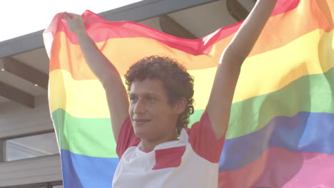 Happy-biracial-man-holding-rainbow-lgbt-flag-on-balcony-at-home,-slow-motion