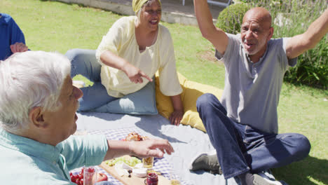 Two-happy-diverse-senior-men-talking-at-a-picnic-with-friends-in-sunny-garden,-slow-motion