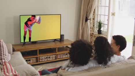 Biracial-family-watching-tv-with-caucasian-male-rugby-player-jumping-with-ball-on-screen