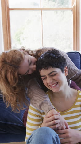 Happy-caucasian-lesbian-couple-sitting,embracing,kissing-and-smiling-in-sunny-living-room