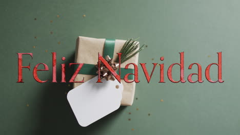 Fekiz-navidad-text-in-red-over-christmas-gift-with-blank-tag-on-green-background