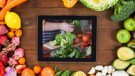 Tablet-with-woman-making-salad-and-vegetables-on-wooden-background