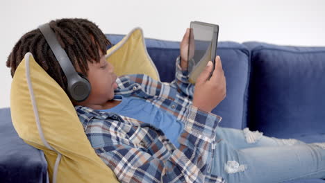 Niño-Afroamericano-Con-Auriculares-Usando-Tableta-Tumbado-En-El-Sofá-En-Casa,-Cámara-Lenta