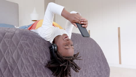 Happy-african-american-boy-wearing-headphones-lying-on-bed-using-smartphone-at-home,-slow-motion