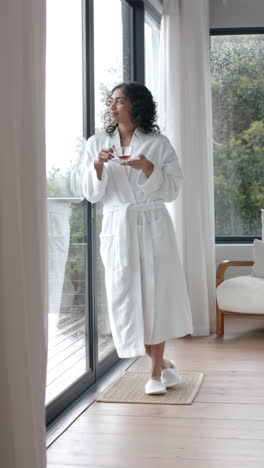 Vertical-video-of-biracial-woman-wearing-bathrobe-drinking-tea-at-home,-slow-motion