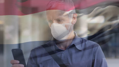 Animation-Eines-Wehenden-Niederländischen-Flaggenschwenkers-über-Einem-Kaukasischen-Mann-Mit-Gesichtsmaske,-Der-Sein-Smartphone-Auf-Der-Straße-Benutzt