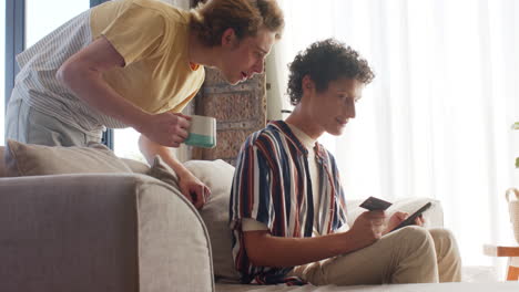 Happy-diverse-gay-male-couple-using-smartphone-for-online-shopping-on-sofa-at-home,-slow-motion