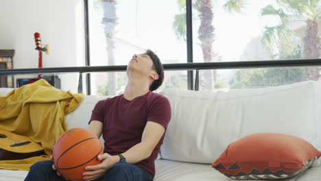 Asiatischer-Männlicher-Teenager,-Der-Mit-Basketball-Fernsieht-Und-Im-Wohnzimmer-Sitzt