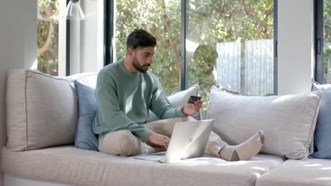 Biracial-man-sitting-on-sofa-using-laptop-for-online-shopping-at-home,-slow-motion